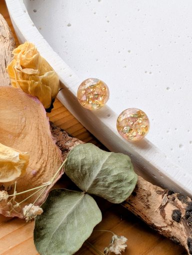 Zwei transparente Ohrstecker mit glitzernden Elementen, umgeben von getrockneten Blumen.